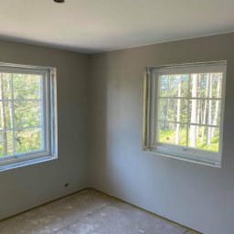Mateusz Górski - Pokój po malowaniu ścian na szaro, z dwoma oknami z widokiem na las. Podłoga przygotowana do dalszych prac wykończeniowych, widoczne zabezpieczenia okien.