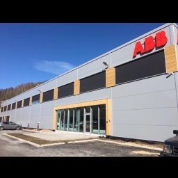 Pol-Nor Invest - Szary budynek biurowy z czerwonym logo ABB, drewnianymi akcentami i szklanymi drzwiami wejściowymi. Błękitne niebo w tle, samochody na parkingu.