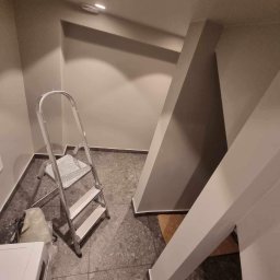 Mateusz Górski - Wnętrze po malowaniu: szara ściana, drabina malarska, widoczne pomalowane elementy konstrukcyjne, szara podłoga. Świeżo odmalowane pomieszczenie.