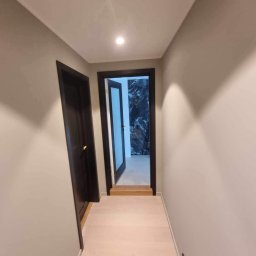 Mateusz Górski - Korytarz po remoncie z jasną podłogą, szarymi ścianami i ciemnymi drzwiami. W tle widoczna ściana z marmurowym wzorem. Oświetlenie punktowe.