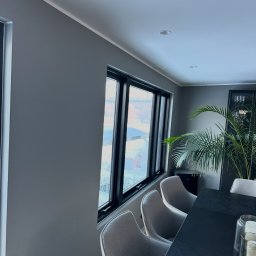 Mateusz Górski - Szare ściany w nowoczesnym wnętrzu z dużymi oknami i czarną ramą, widok na zimowy krajobraz, stół z krzesłami i rośliną doniczkową w tle.