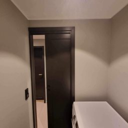 Mateusz Górski - Ciemne, przesuwne drzwi z widokiem na kolejne pomieszczenie, szare ściany i fragment białej pralki. Minimalistyczne wnętrze po remoncie.