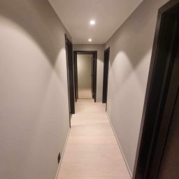 Mateusz Górski - Długi korytarz po remoncie z jasną podłogą i szarymi ścianami. Czarne ramy drzwi i punktowe oświetlenie sufitowe. Widok w głąb, perspektywa zbieżna.