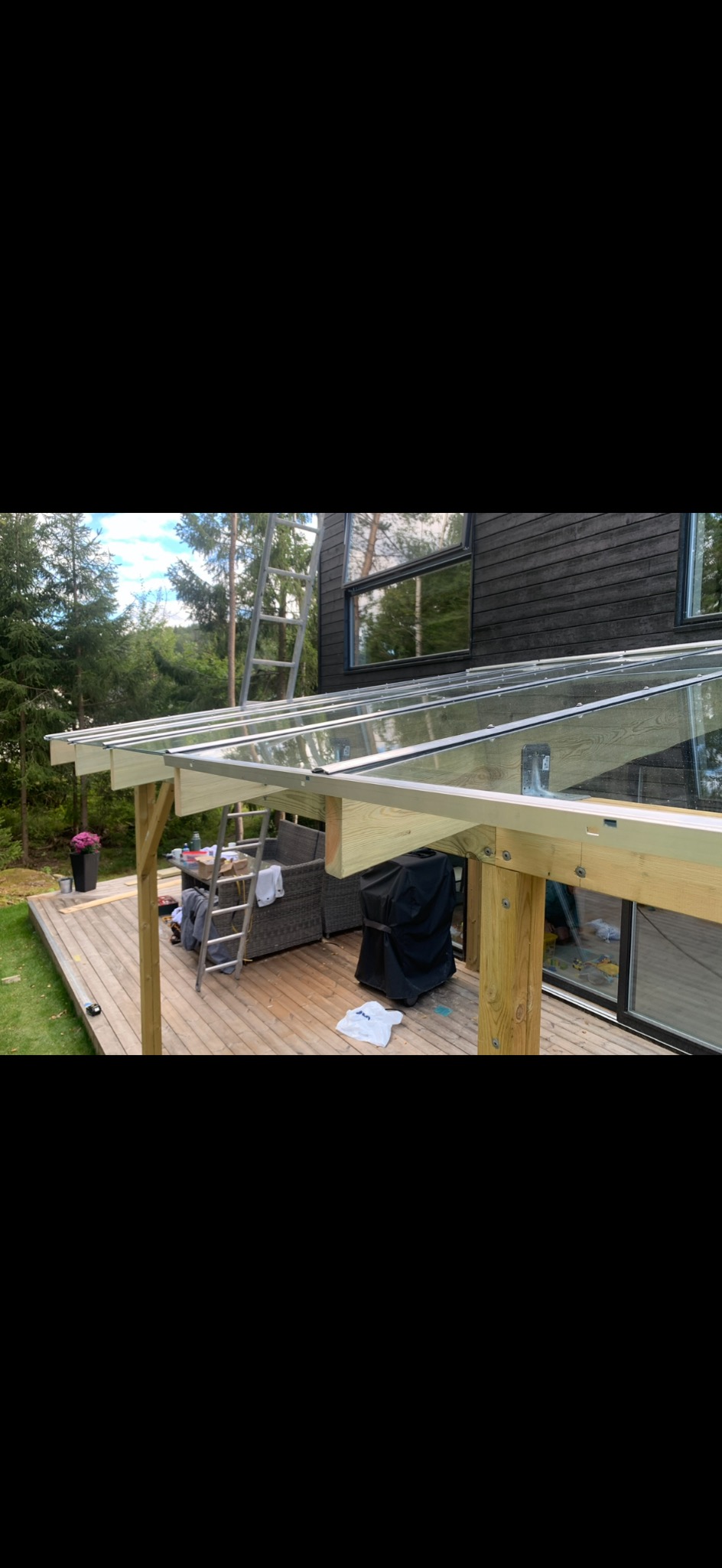 Zadaszenie tarasu z przezroczystego szkła i drewnianej konstrukcji, widok z zewnątrz, drabina oparta o konstrukcję, grill pod przykryciem na drewnianym tarasie.