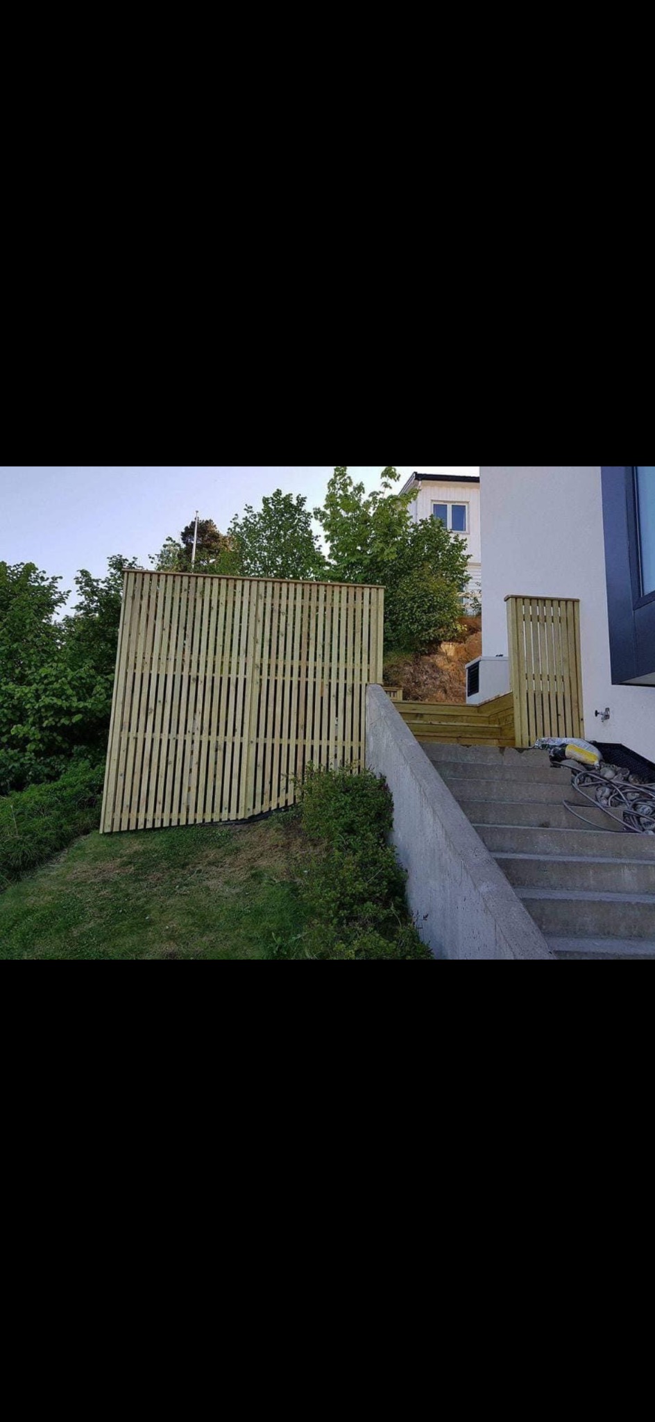 Nowe, jasne, drewniane ogrodzenie panelowe na nierównym terenie, z betonowymi schodami prowadzącymi do domu. Widoczne drzewa i krzewy w tle.