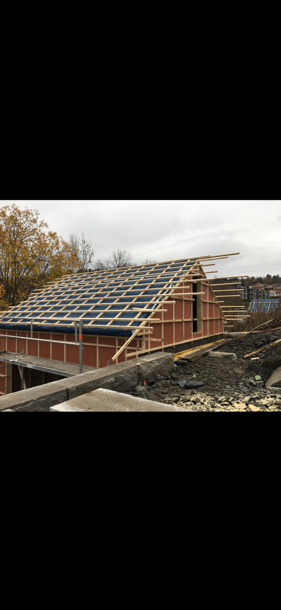 Konstrukcja dachu w trakcie budowy, widoczne drewniane elementy szkieletu i niebieska membrana dachowa. W tle zabudowa mieszkalna i drzewa.