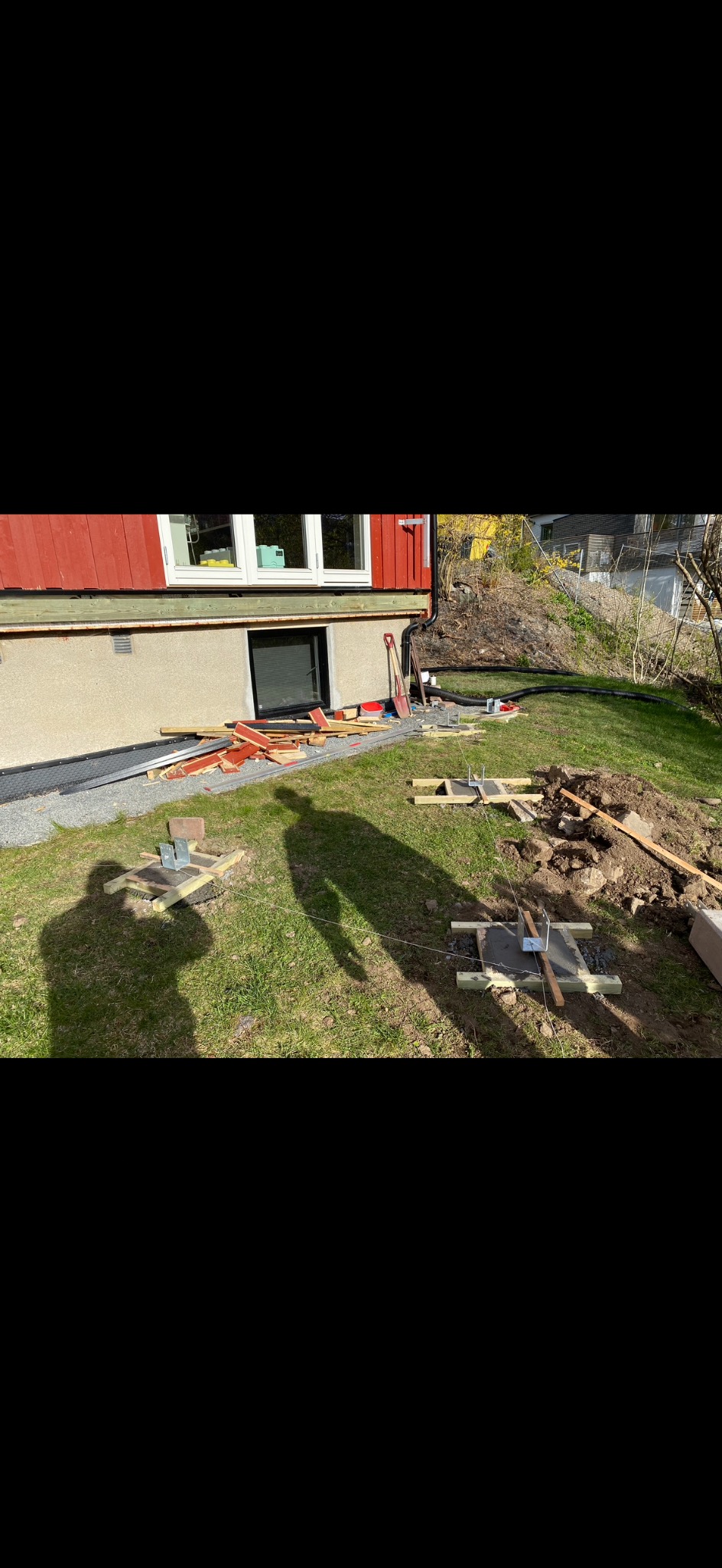 Fundamenty pod budynek w trakcie budowy. Widoczne drewniane szalunki, beton, metalowe elementy mocujące i cień osoby na trawie. Prace ziemne w toku.