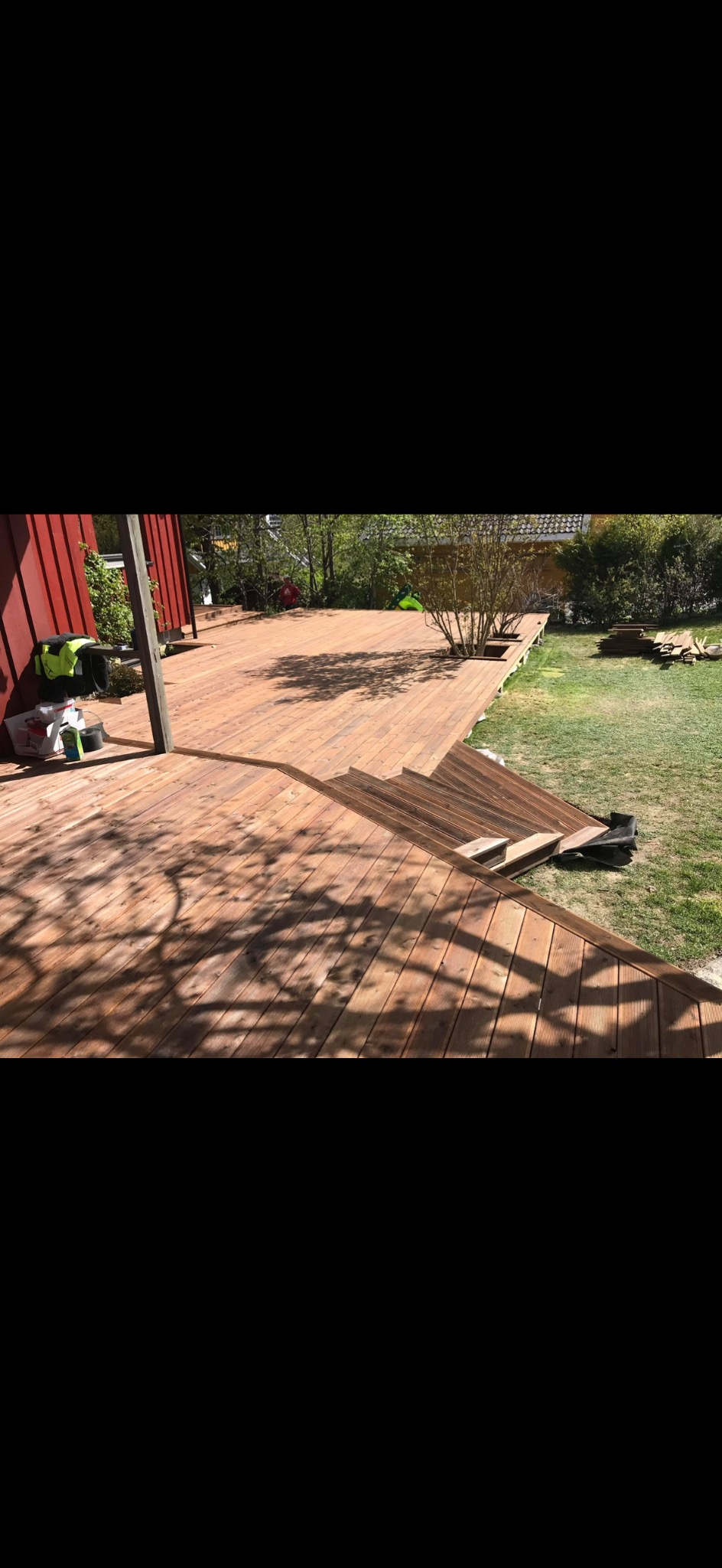 Drewniany taras z niskimi schodami, otoczony zielenią. Widoczne narzędzia i materiały budowlane. Cień drzewa na deskach. Ujęcie z boku, w dziennym świetle.