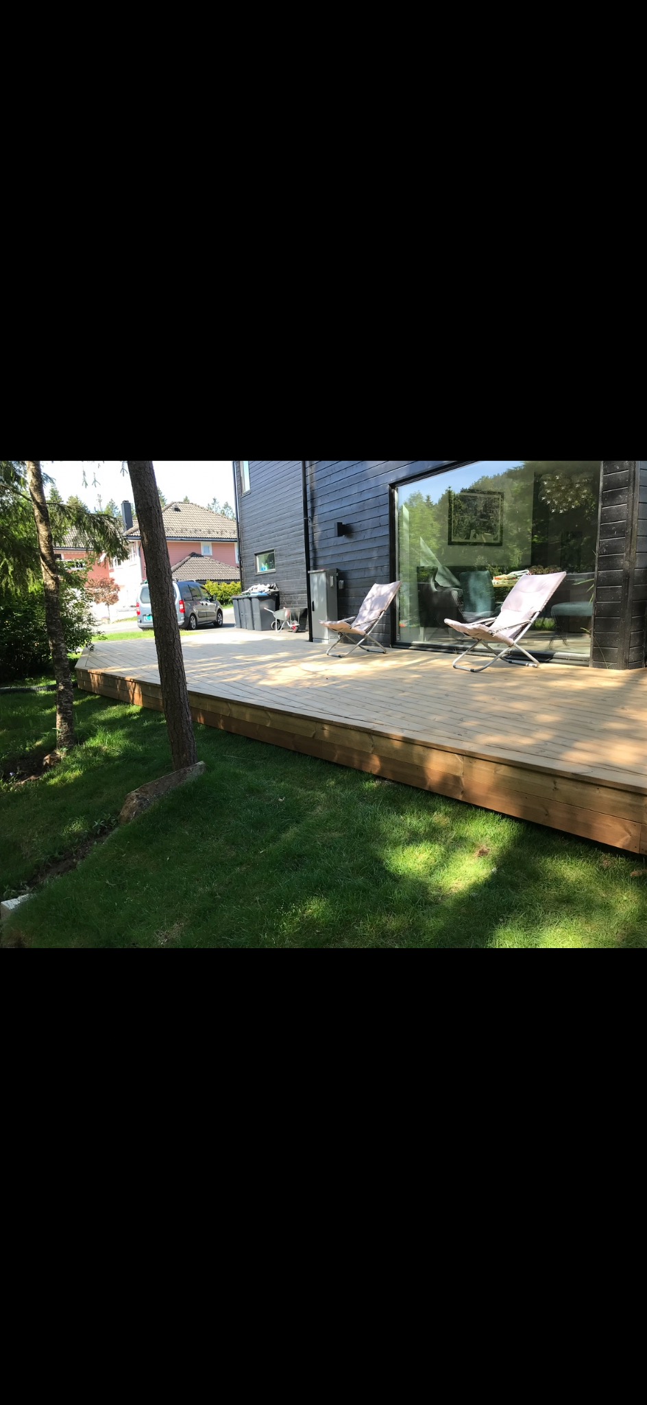 Nowoczesny taras z jasnego drewna przy czarnym domu, z dwoma leżakami i dużym oknem. Widok na zadbany trawnik i drzewa.