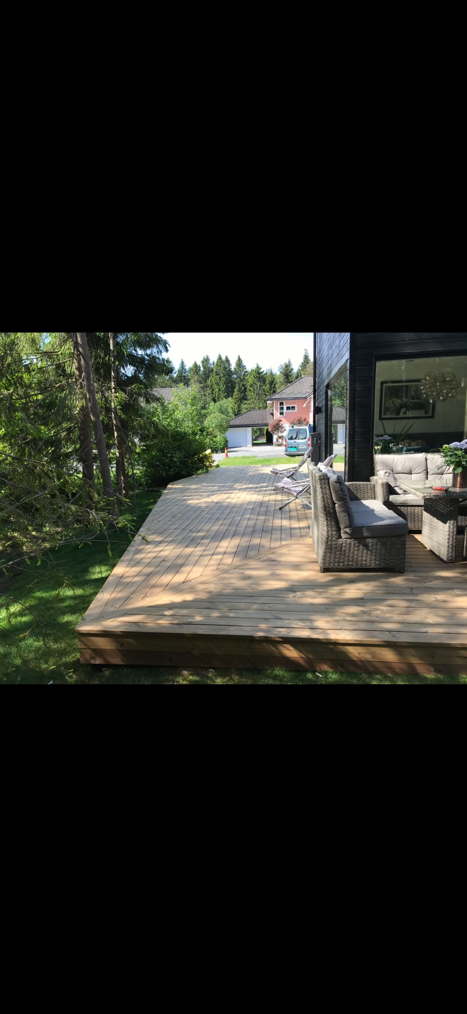 Jasna, drewniana podłoga tarasu z meblami ogrodowymi w odcieniach szarości, otoczona zielenią trawnika i drzew. W tle dom z dużym oknem.