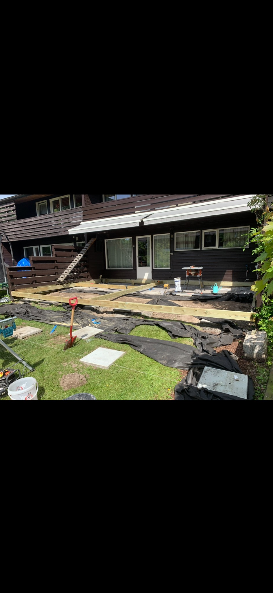 Budowa drewnianego tarasu przy domu. Widoczne fundamenty, częściowo ułożona konstrukcja z belek, narzędzia i materiały budowlane na trawniku.