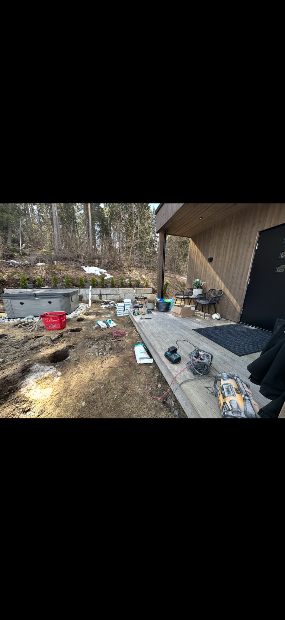 Montaż drewnianego tarasu przy domu z jacuzzi w tle. Widoczne narzędzia, materiały budowlane i wykopane dołki pod konstrukcję. Prace w toku, słoneczny dzień.