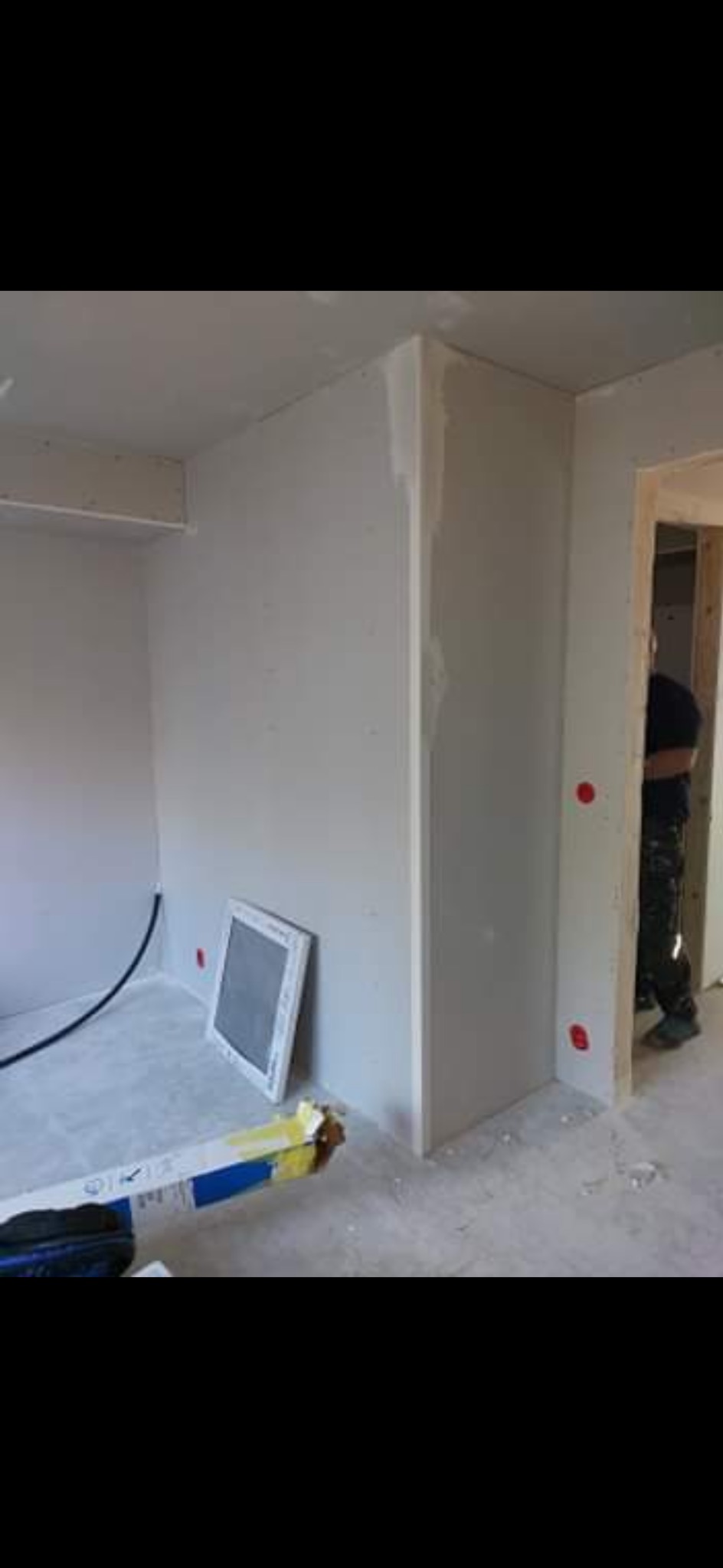 Wnętrze w trakcie prac wykończeniowych: ściany z płyt karton-gips, widoczne łączenia, fragment okna i narzędzia. W tle sylwetka osoby.