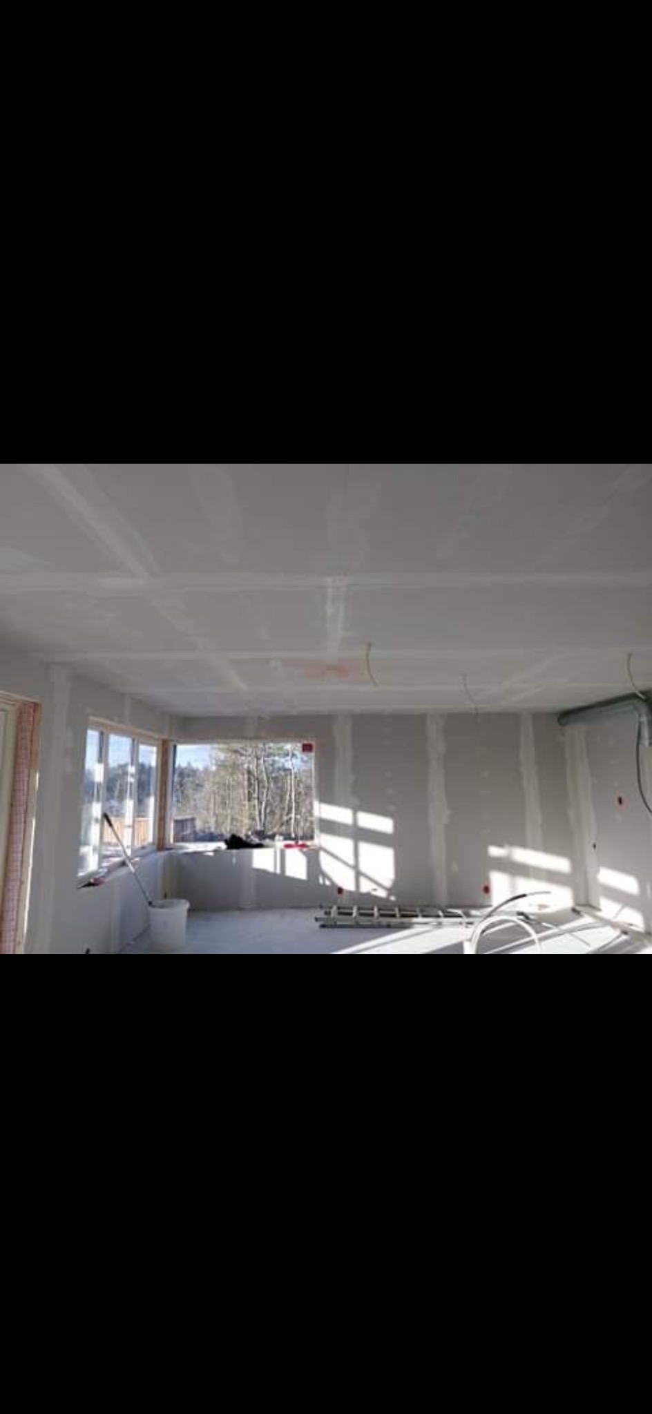 Pokój w trakcie prac wykończeniowych: ściany i sufit pokryte płytami kartonowo-gipsowymi, widoczne okna z naturalnym światłem, drabina i wiadro na podłodze.