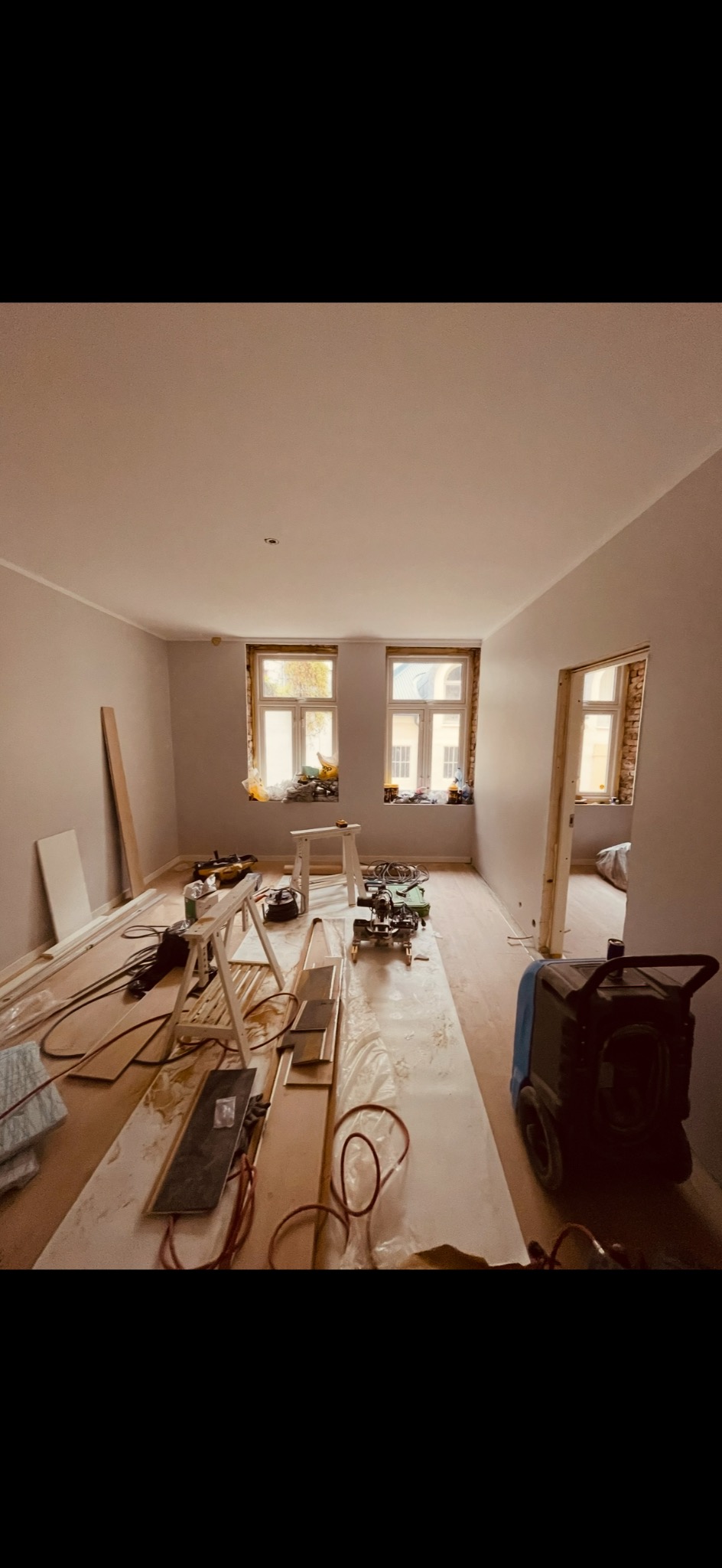 Jasne wnętrze w trakcie prac wykończeniowych: świeżo pomalowane ściany, ułożona podłoga, narzędzia i materiały budowlane rozłożone na podłodze, widok na okna.