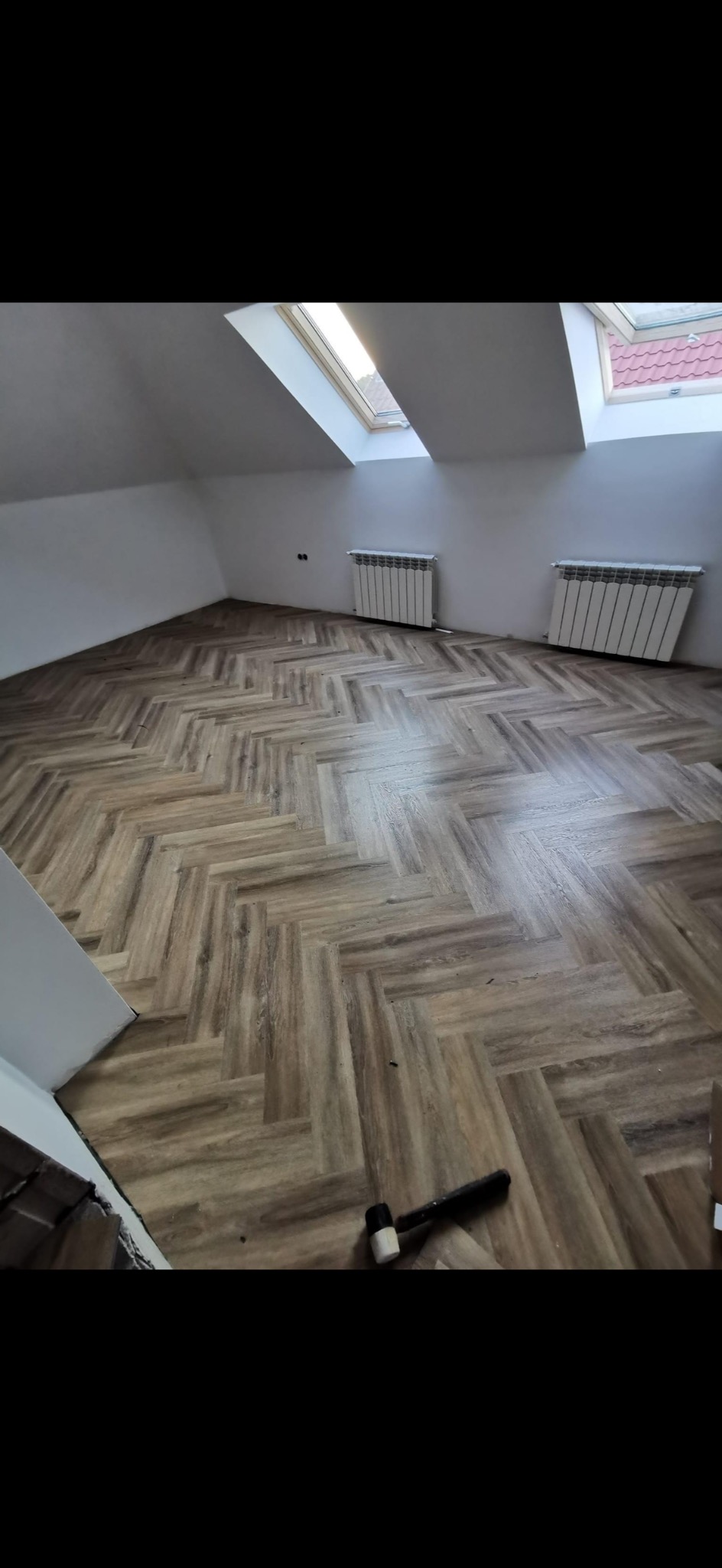 Jasne panele ułożone w jodełkę w pokoju na poddaszu z oknami dachowymi i grzejnikami. Gumowy młotek leży na podłodze, sugerując trwający montaż.
