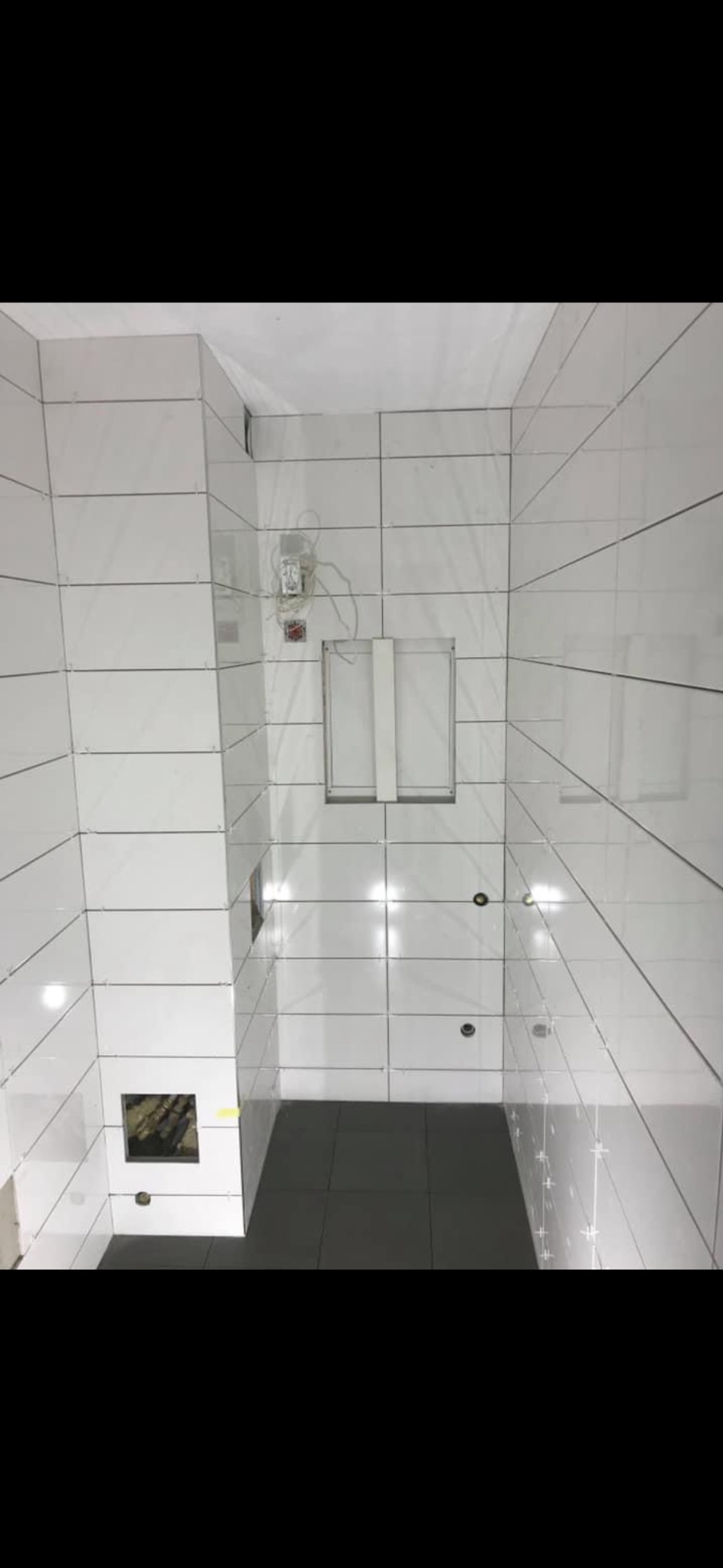 Białe, prostokątne płytki na ścianach i szare na podłodze w trakcie remontu łazienki. Widoczne otwory na instalacje i wystające kable. Ujęcie z góry.
