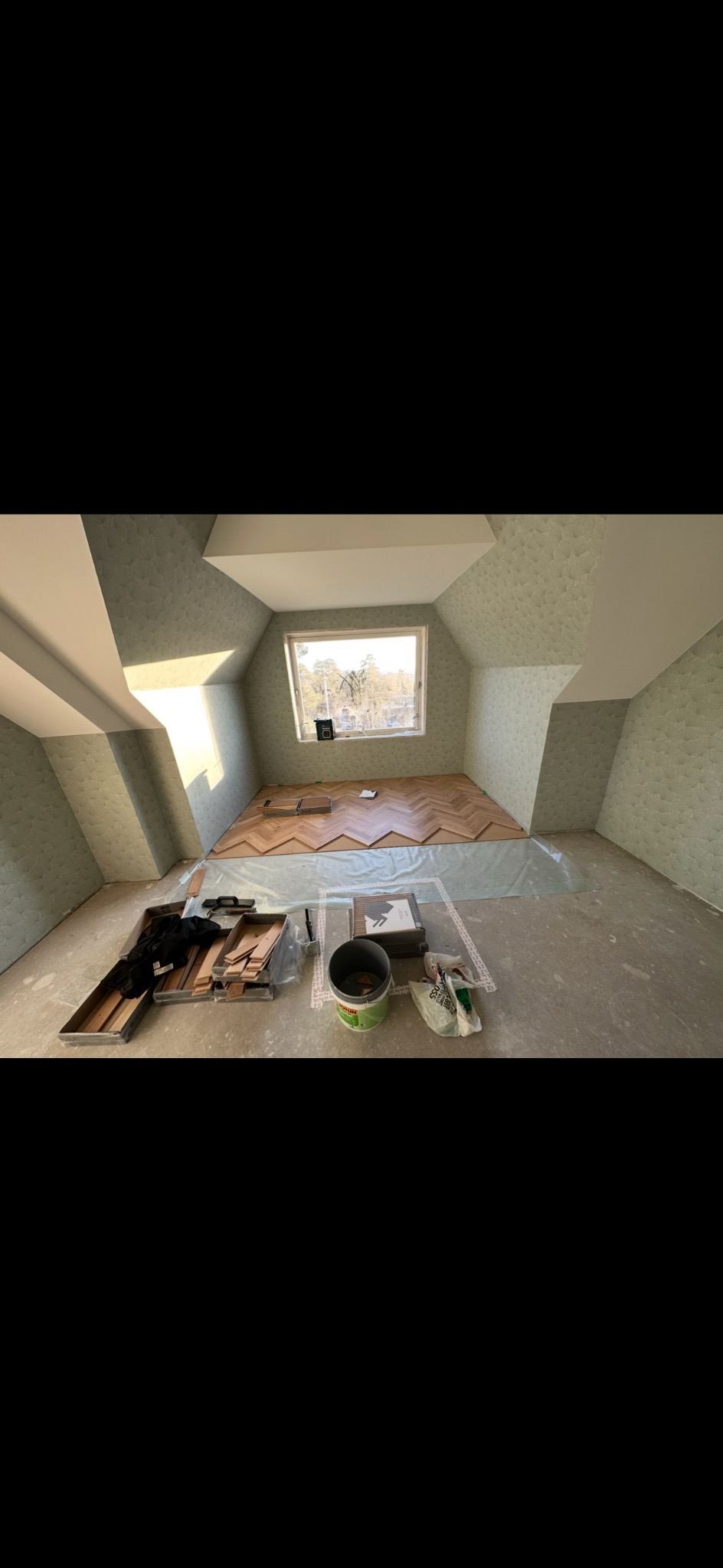 Układanie parkietu w jodełkę w pokoju na poddaszu. Widoczne narzędzia i materiały przygotowane do montażu. Część podłogi zabezpieczona folią. Ściany w jasnym kolorze.