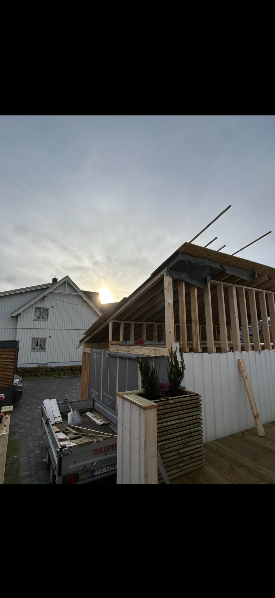 Konstrukcja drewniana w trakcie budowy, widoczny szkielet dachu i ścian. W tle biały dom i przyczepa z materiałami budowlanymi. Słońce przebija się przez chmury.