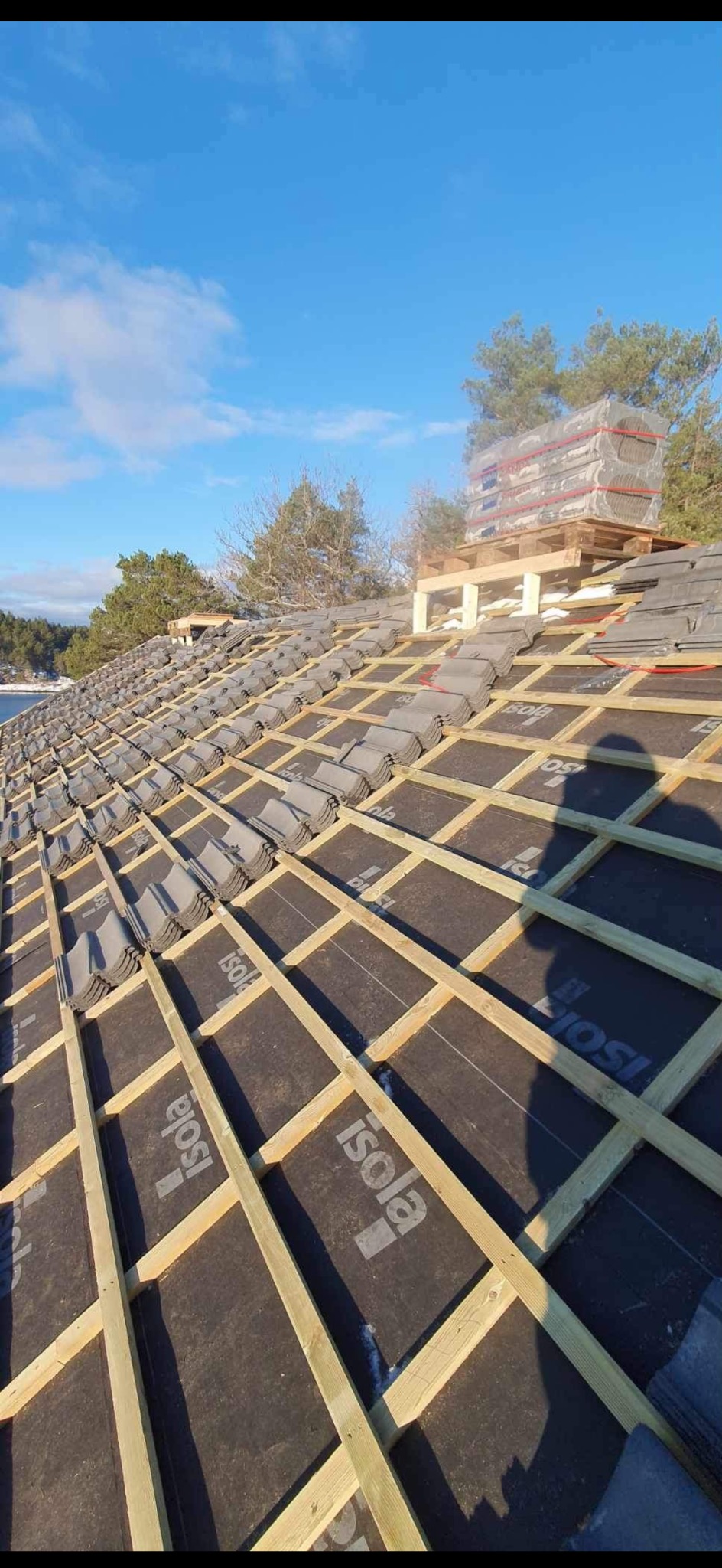 Montaż dachówki ceramicznej na łatach z folią izolacyjną. Częściowo pokryty dach z paletą dachówek w tle, błękitne niebo.