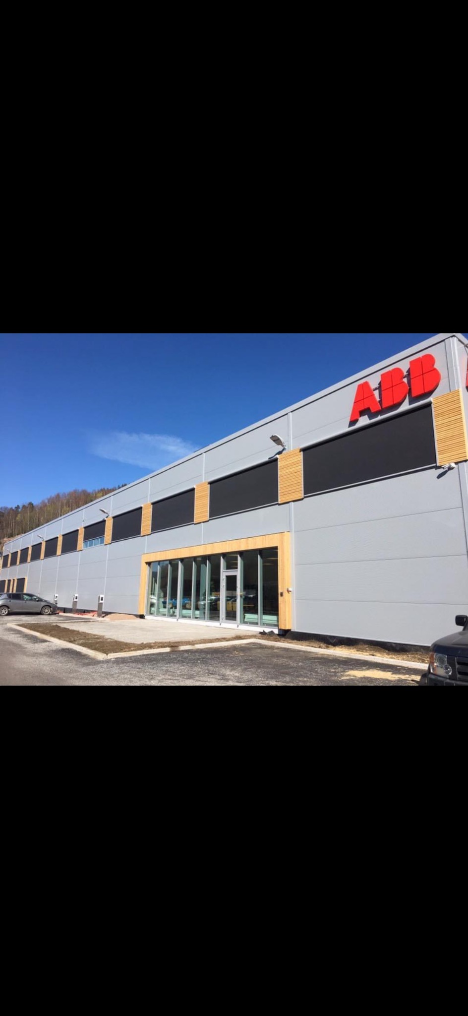 Szary budynek biurowy z czerwonym logo ABB, drewnianymi akcentami i szklanymi drzwiami wejściowymi. Błękitne niebo w tle, samochody na parkingu.