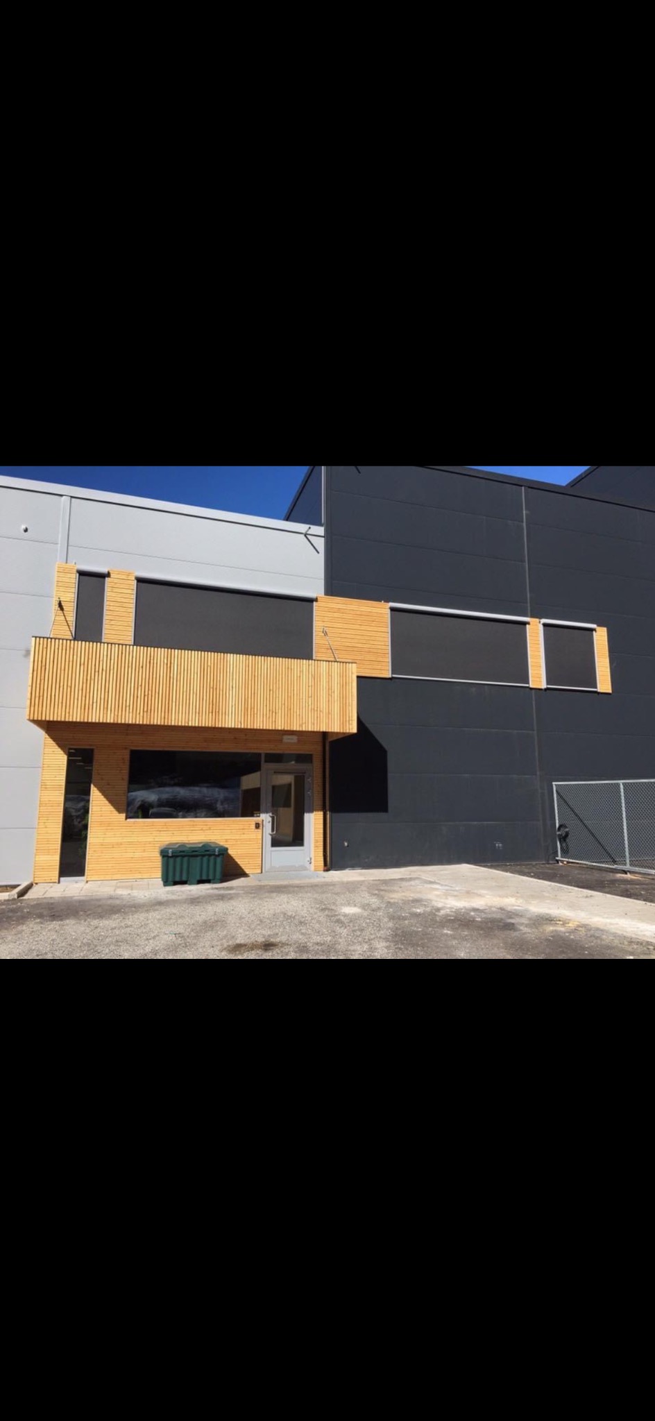 Nowoczesny budynek biurowy z elementami drewnianymi na elewacji, szarymi ścianami i przeszklonym wejściem. Widoczny fragment chodnika i zielony pojemnik na zewnątrz.