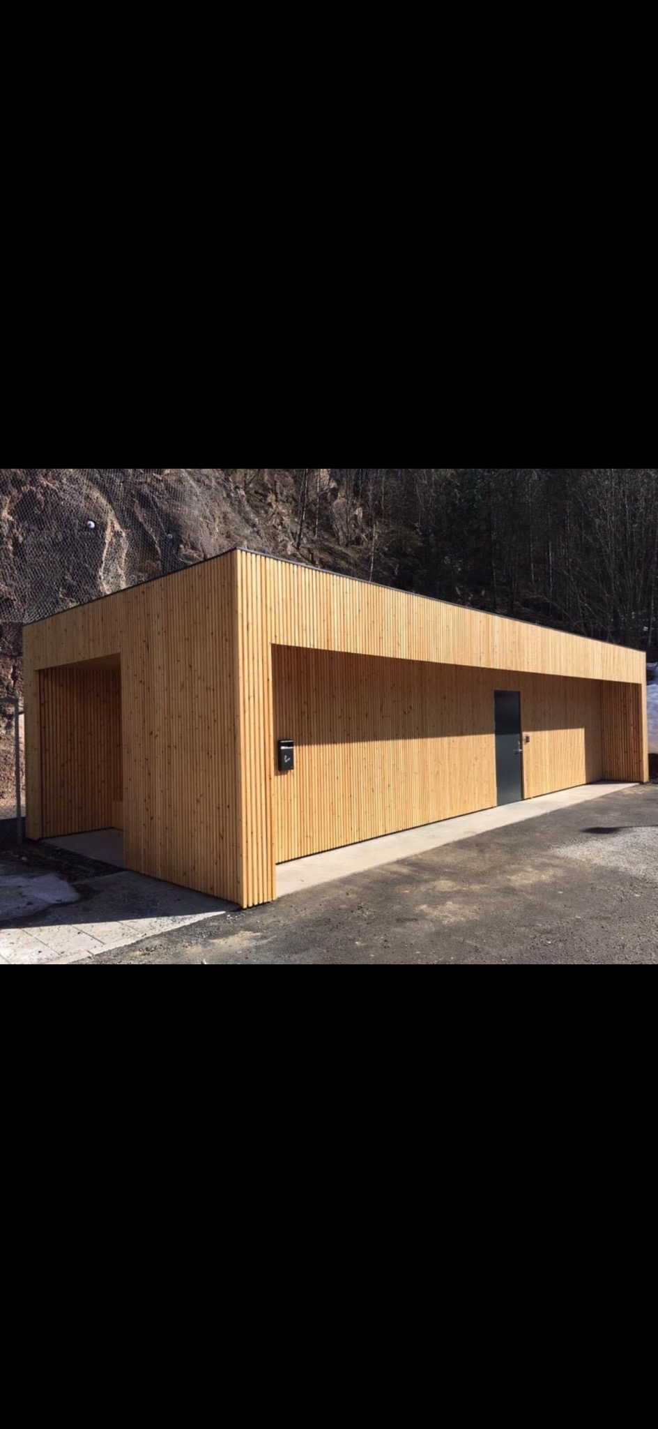 Nowoczesny budynek z jasnego drewna z czarnymi akcentami, wkomponowany w naturalne otoczenie skalistej góry i drzew. Minimalistyczny design z wyraźnymi liniami.