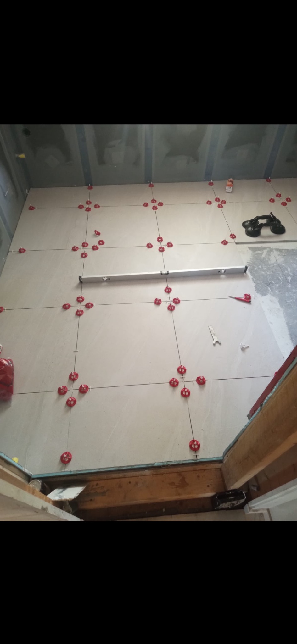 Układanie jasnych płytek podłogowych z użyciem krzyżyków dystansowych i poziomicy. Widoczne narzędzia glazurnicze i przygotowane podłoże.