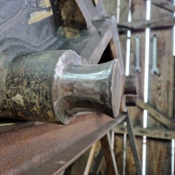 KAMIL SZCZECHURA - Wyszlifowany element metalowy na tle drewnianej konstrukcji z narzędziami. Widoczne ślady obróbki i rdzy. Surowe oświetlenie podkreśla detale.