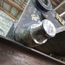 KAMIL SZCZECHURA - Szlifowany sworzeń mocowania łyżki koparki po naprawie, leżący na stalowym profilu. Widoczne ślady wcześniejszego zużycia i obróbki. Surowe otoczenie warsztatowe.