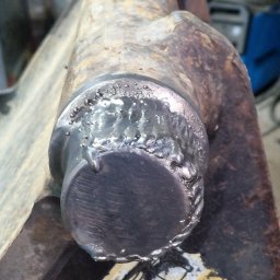 KAMIL SZCZECHURA - Spoina na metalowym elemencie, widoczne ślady obróbki termicznej i stopiony metal. Niejednolita powierzchnia z widocznymi nierównościami i przebarwieniami.