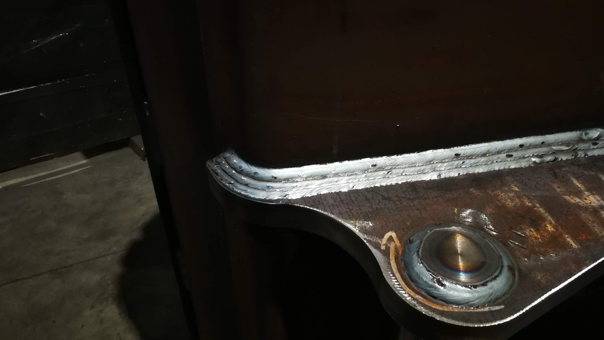 Spoina spawalnicza na metalowej konstrukcji, widoczne ślady obróbki i charakterystyczne przebarwienia termiczne. Detal z bliska, ciemne tło.