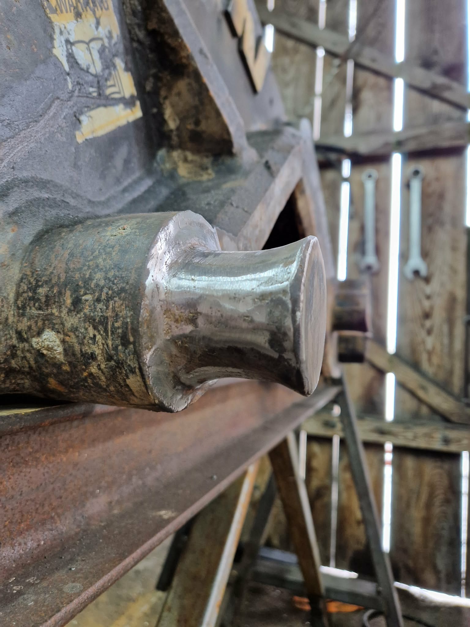 Wyszlifowany element metalowy na tle drewnianej konstrukcji z narzędziami. Widoczne ślady obróbki i rdzy. Surowe oświetlenie podkreśla detale.