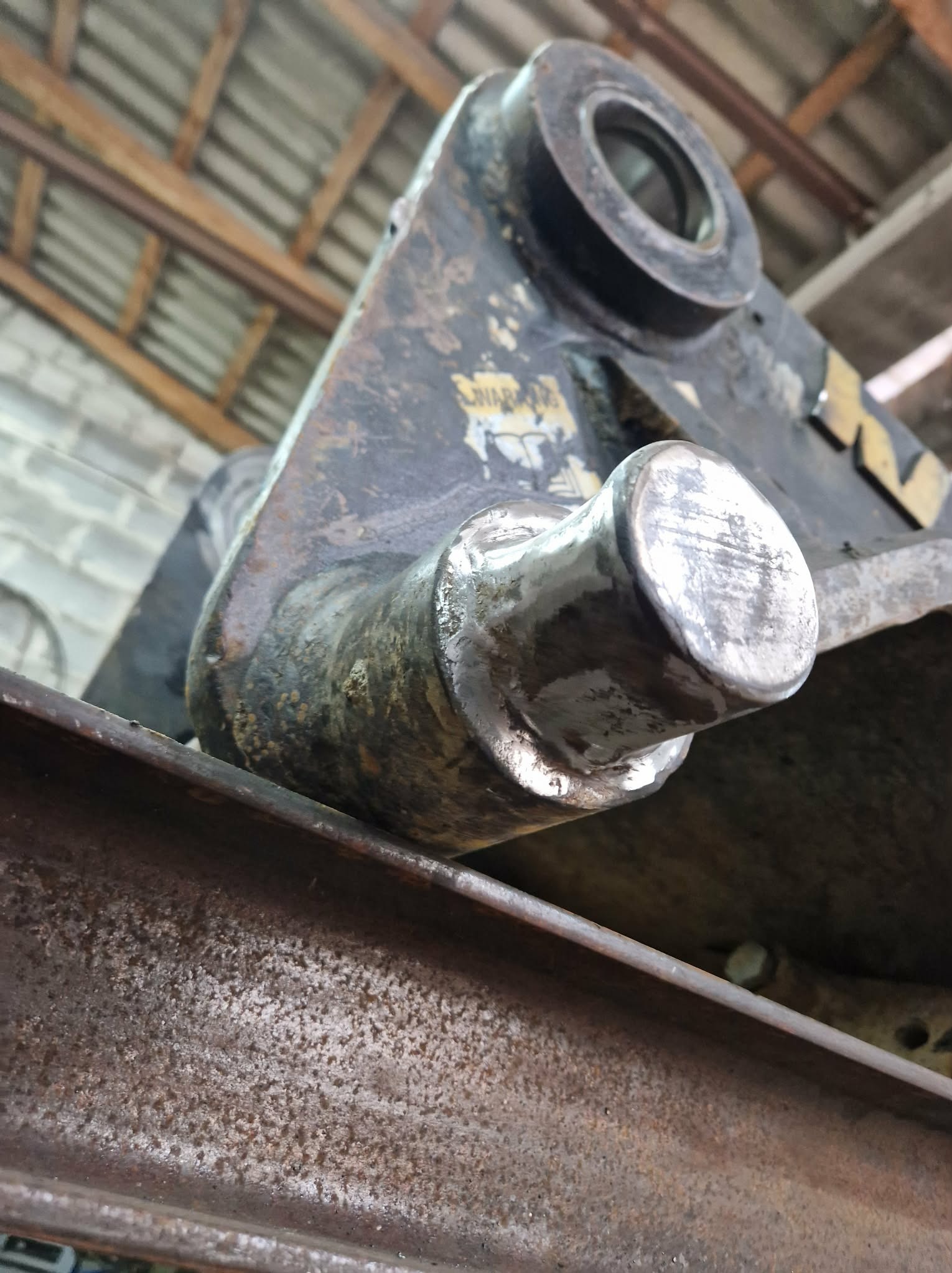 Szlifowany sworzeń mocowania łyżki koparki po naprawie, leżący na stalowym profilu. Widoczne ślady wcześniejszego zużycia i obróbki. Surowe otoczenie warsztatowe.