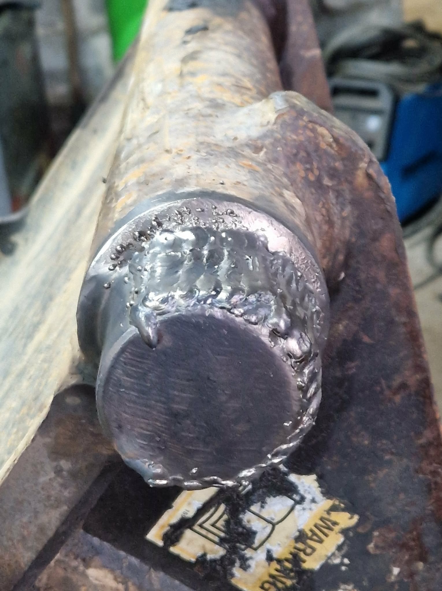 Spoina na metalowym elemencie, widoczne ślady obróbki termicznej i stopiony metal. Niejednolita powierzchnia z widocznymi nierównościami i przebarwieniami.