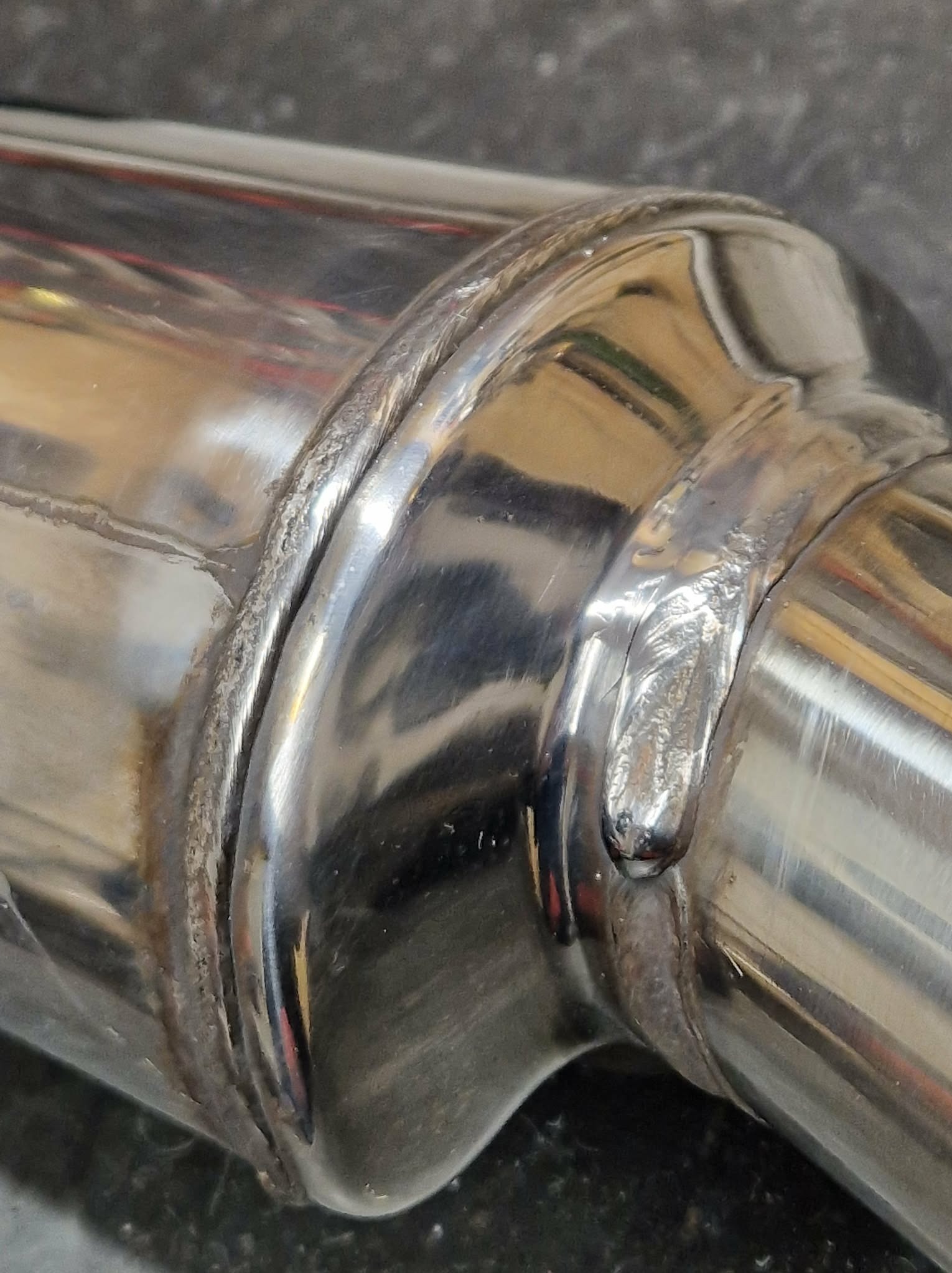 Błyszczące, metalowe rury połączone precyzyjnym spawem, widoczne detale spoiny i odbicia światła na gładkiej powierzchni metalu. Makro zbliżenie na łączenie.