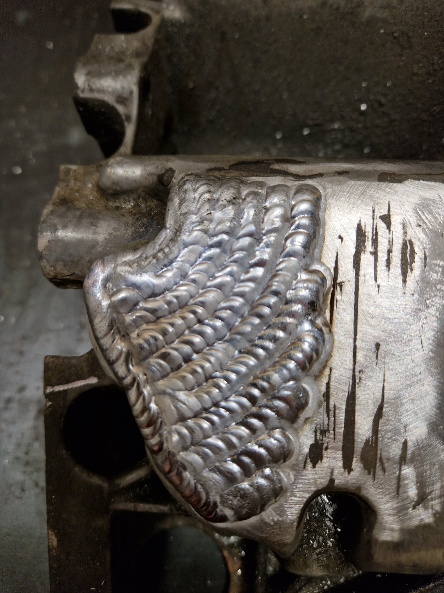 Precyzyjne spawy na metalowym elemencie, układające się w charakterystyczny wzór. Widoczne ślady obróbki i surowość materiału.