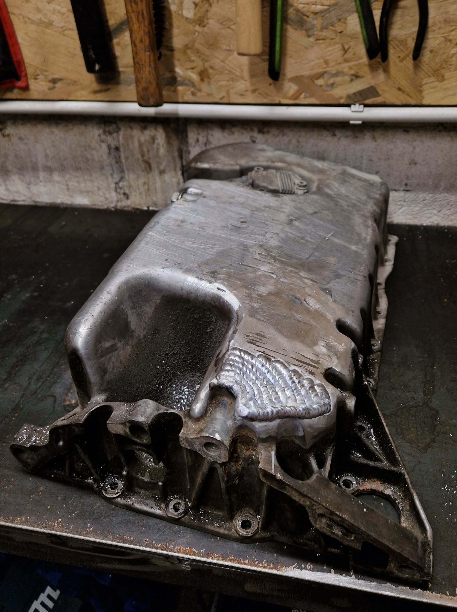 Naprawiona, aluminiowa miska olejowa po spawaniu, widoczne spawy. W tle warsztat z narzędziami.