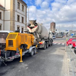 Grupa Almar Serwis Sp. z o.o. - Żółta pompa do betonu MECBO i betonomieszarka Liebherr na ulicy, w tle budynek z czerwonym dachem i wieża. Dzień, pochmurne niebo.