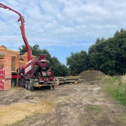 Grupa Almar Serwis Sp. z o.o. - Czerwono-biała betonomieszarka z wysięgnikiem na placu budowy, obok palety z cegłami Porotherm. W tle zielone drzewa i fragment wznoszonej konstrukcji.