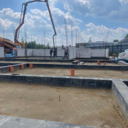 Grupa Almar Serwis Sp. z o.o. - Wylewanie fundamentów pod budynek z użyciem pompy do betonu. Widoczne zbrojenia, bloczki fundamentowe i pracownicy budowlani. Słoneczny dzień, błękitne niebo z chmurami.