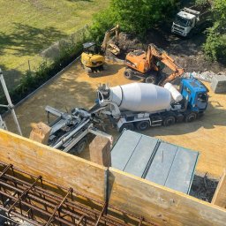 Grupa Almar Serwis Sp. z o.o. - Z góry: betonowóz, pompa do betonu, dwie koparki na placu budowy. Widoczne zbrojenie i szalunki. Słoneczny dzień.