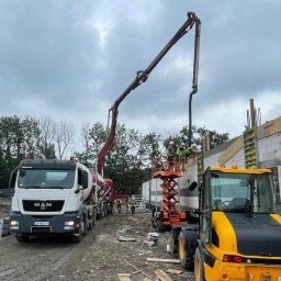 Grupa Almar Serwis Sp. z o.o. - Wylewanie betonu na budowie z użyciem pompy zamontowanej na ciężarówce MAN. Pracownicy w kaskach nadzorują proces z podnośnika. Widoczny żółty traktor i ściany z bloczków.