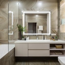 Artem Kobylianskyi - Elegancka łazienka z podświetlanym lustrem, beżowymi płytkami imitującymi marmur i białymi meblami. Uwagę przykuwają dekoracyjne figurki ptaków na blacie.