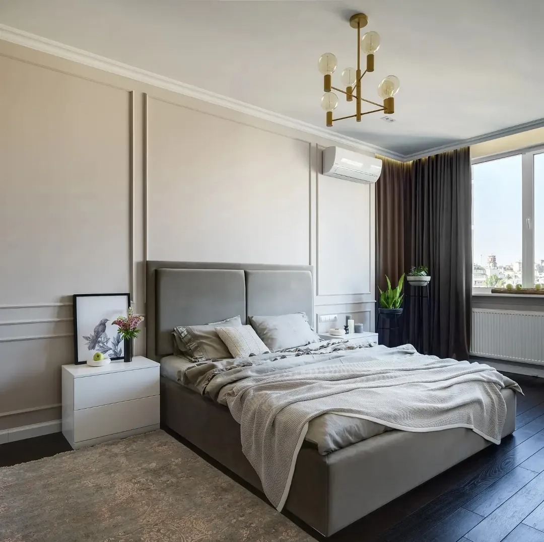 Nowoczesna sypialnia w stonowanych szarościach z ciemną podłogą, listwami dekoracyjnymi na ścianach i złotą lampą sufitową. Widok na miasto przez okno.
