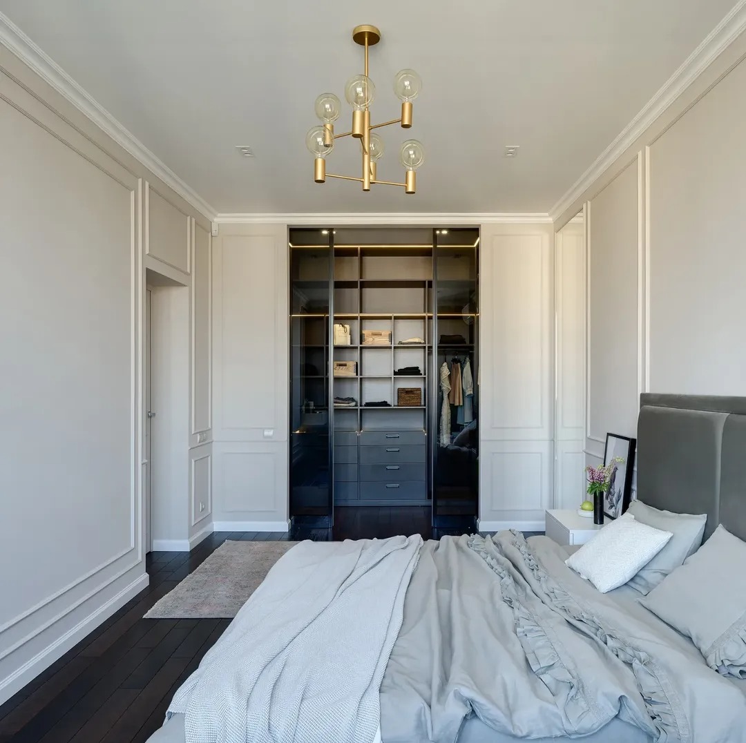Elegancka sypialnia w odcieniach szarości z dużym, przesuwnym lustrzanym garderobą i złotą lampą sufitową. Klasyczne sztukaterie ścienne i ciemna podłoga.