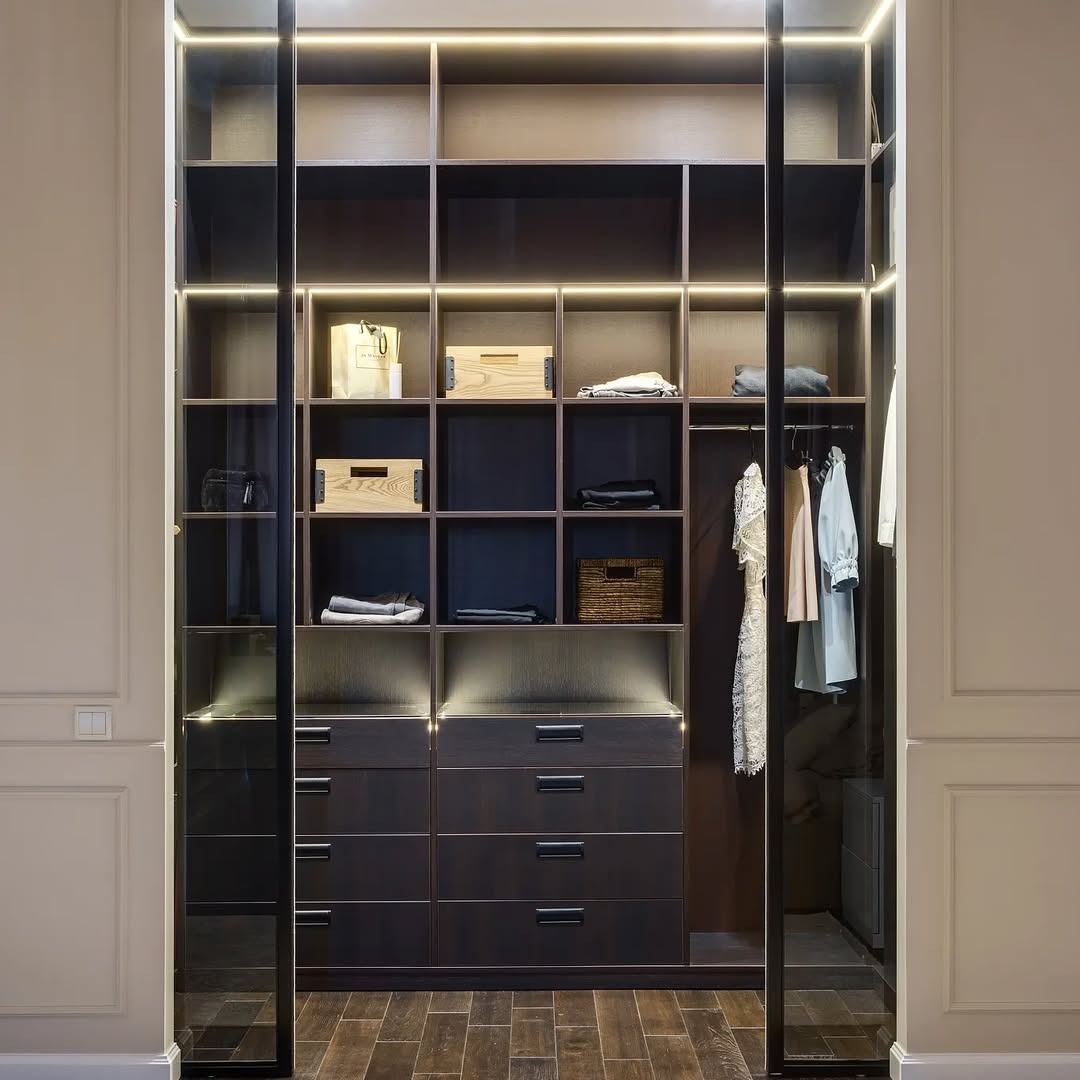 Nowoczesna garderoba z ciemnego drewna, oświetlona LED, z przesuwnymi drzwiami i drewnianą podłogą. Widoczne półki, szuflady i wieszaki z ubraniami.