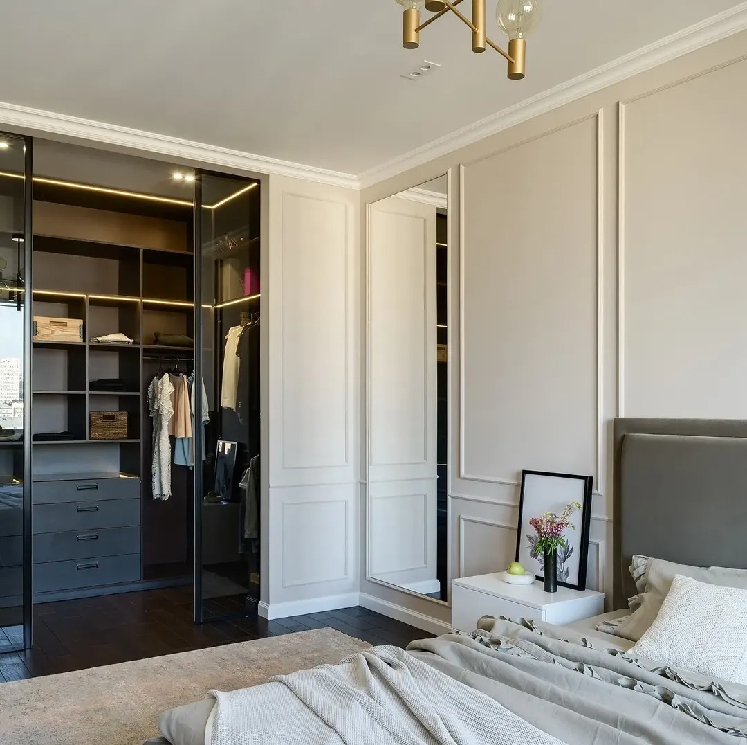 Elegancka sypialnia z dużą szafą wnękową z przesuwnymi, lustrzanymi drzwiami i oświetleniem LED. Klasyczne sztukaterie na ścianach i stonowana kolorystyka.
