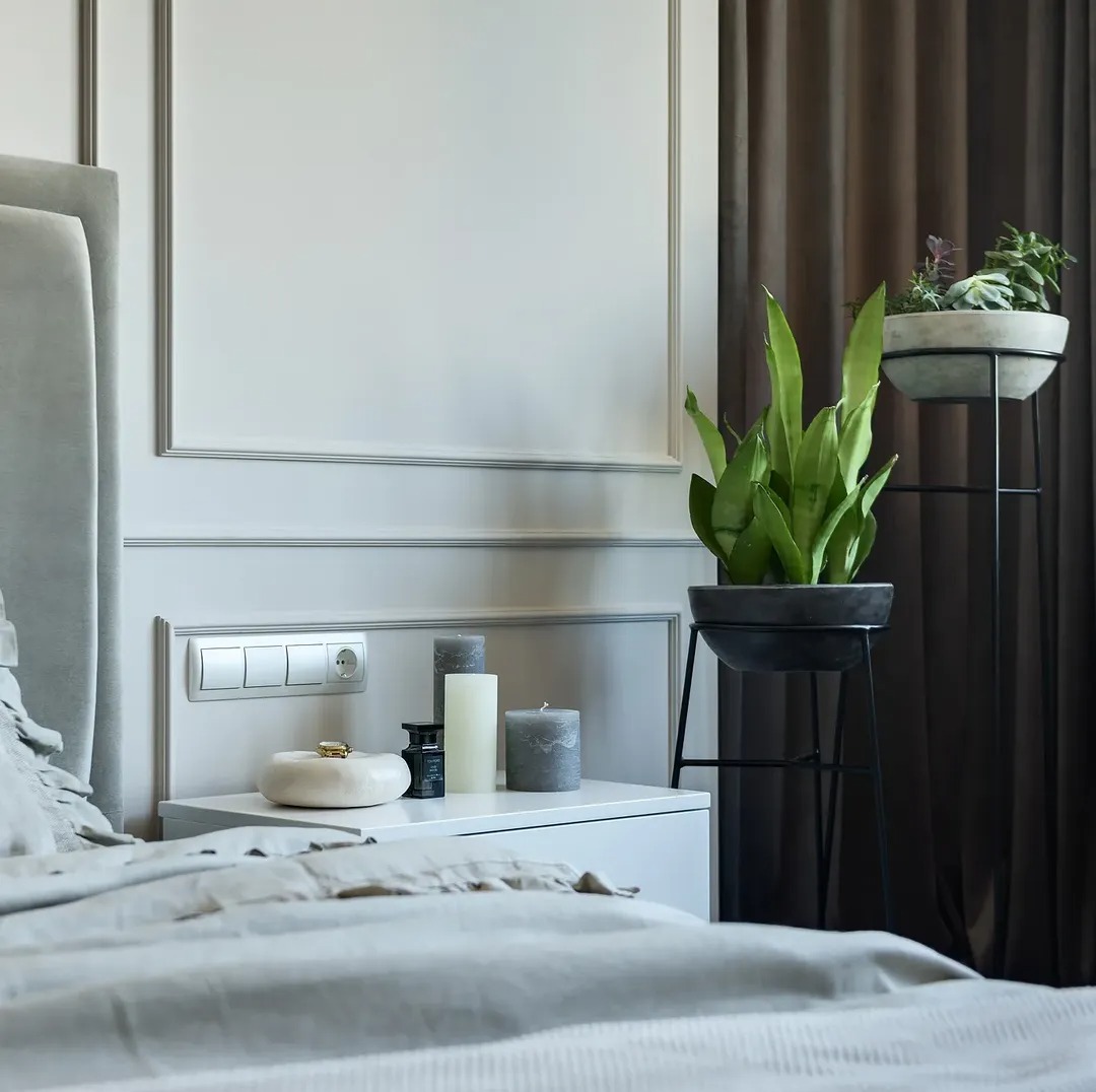 Stylowa sypialnia w odcieniach szarości: łóżko z wysokim wezgłowiem, szafka nocna z dekoracjami (świece, roślina), panel ścienny z gniazdkami i brązowe zasłony.