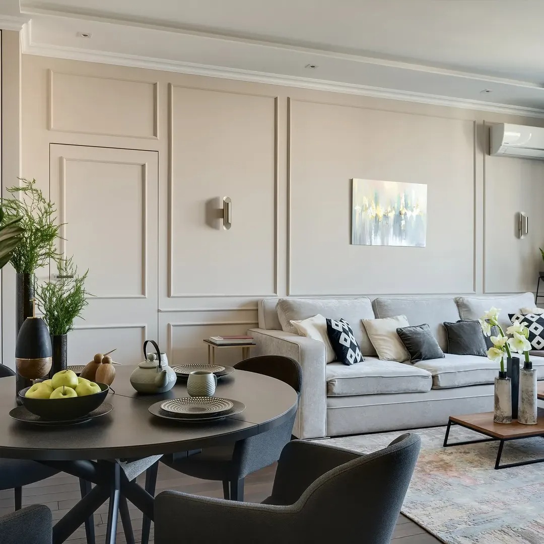 Elegancki salon w beżach: sofa z poduszkami, okrągły stół z zastawą, dekoracyjne listwy ścienne i subtelne oświetlenie. Nowoczesny design z akcentami roślinnymi.